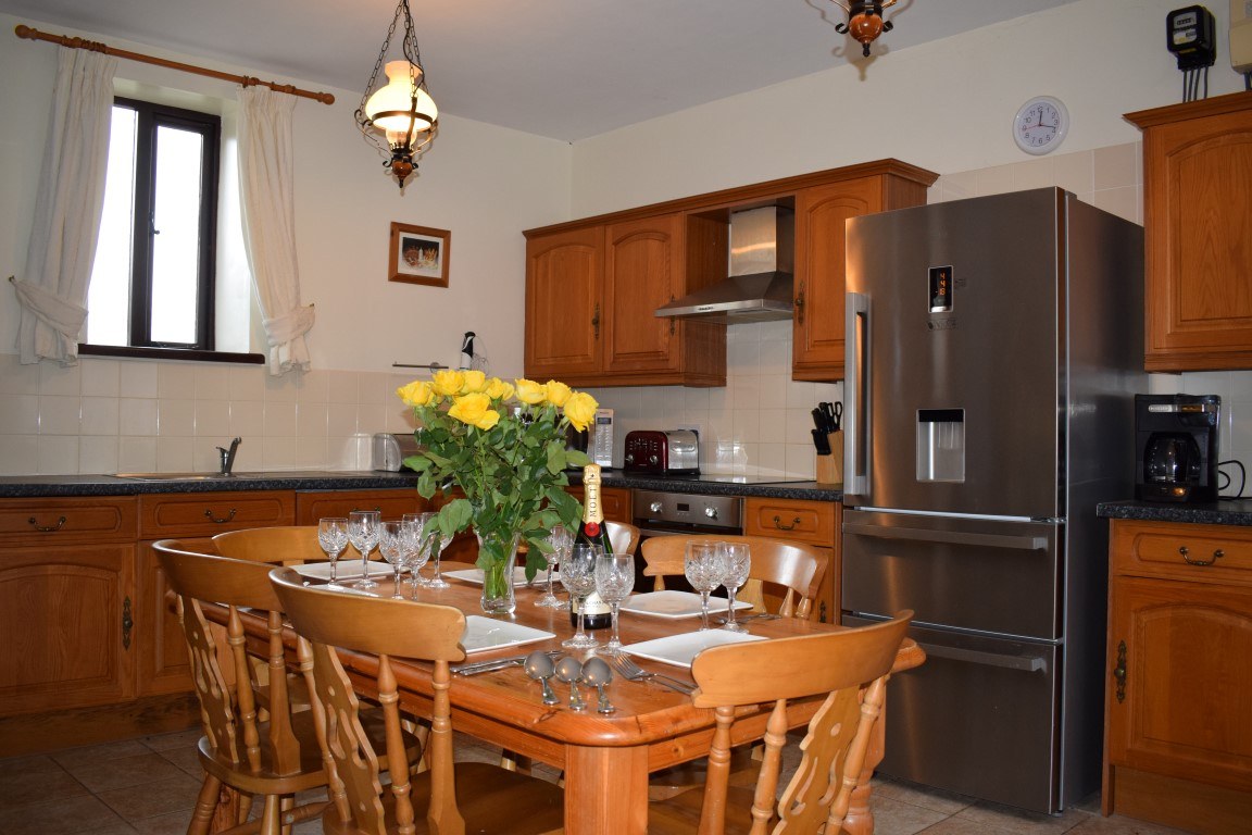 A cottage kitchen with wooden cupboards, a table set with flowers and a fridge freezer