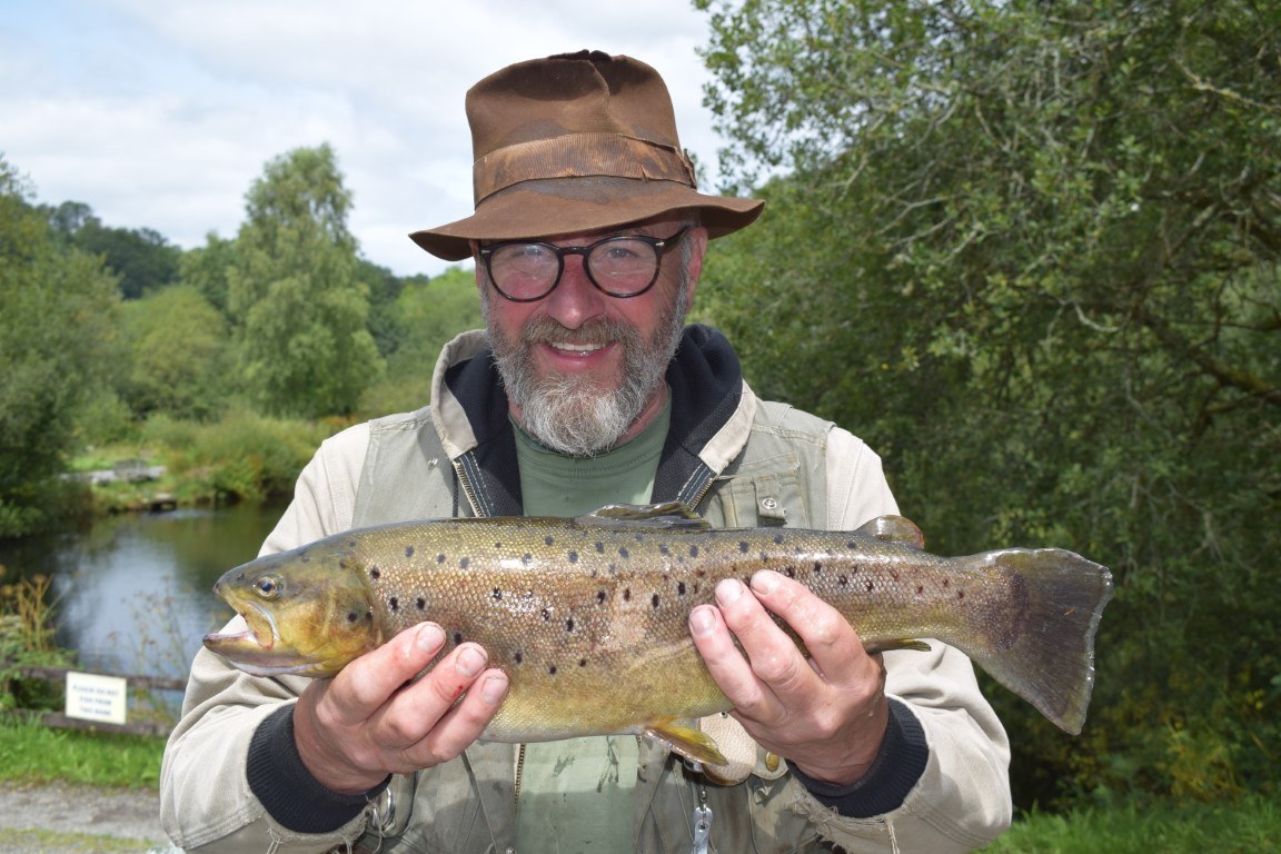 An angler in a wide-brimmed hat holding a brown trout by the lake