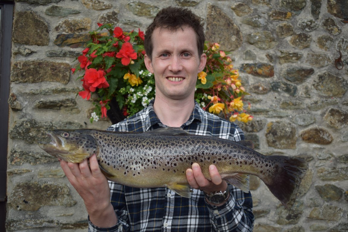 An angler holding a spotted brown trout in front of a stone wall