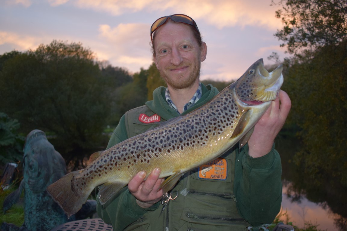 A smiling angler holding a spotted brown trout at dusk